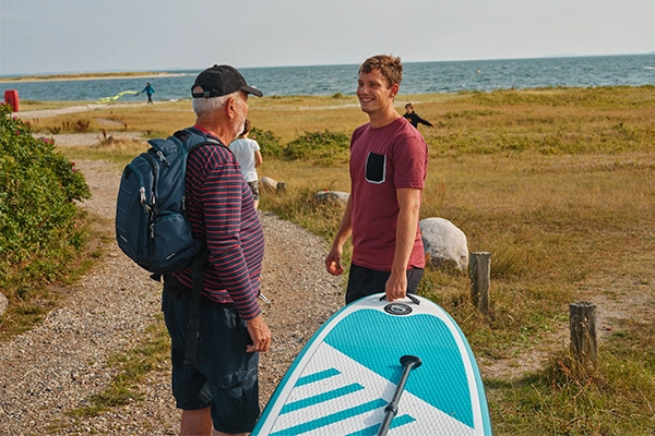 Pensioneret bedstefar og barnebarn med surfbræt og rygsæk ved strand